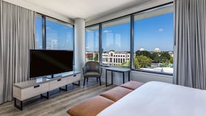 Una habitación de hotel moderna con una cama grande, un televisor sobre un mueble de madera, una silla, una mesita y vistas a la ciudad a través de amplias ventanas.