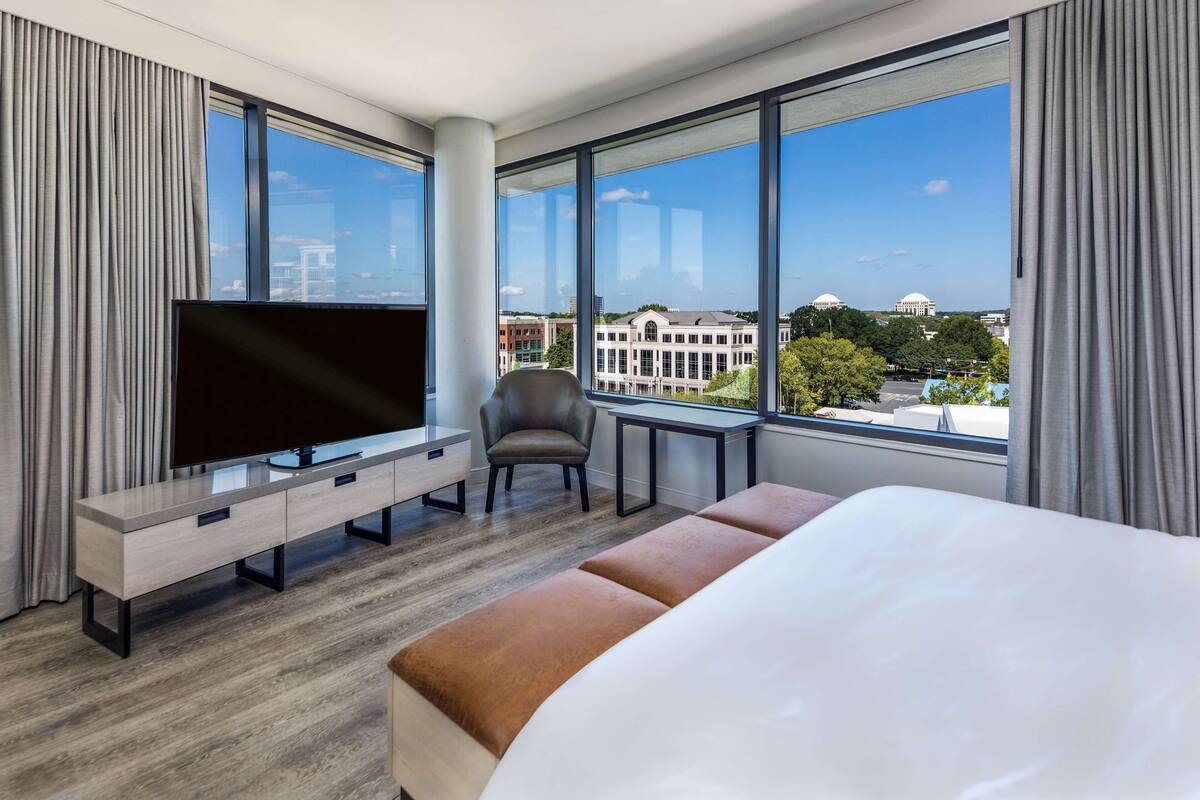 A modern hotel room with a large bed, a TV on a wooden console, a chair, a small table, and a view of the city through large windows.