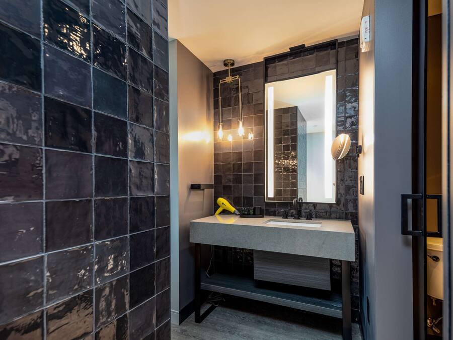 Un baño moderno con paredes de azulejos oscuros, un lavamanos blanco y un espejo con una lámpara sobre él.