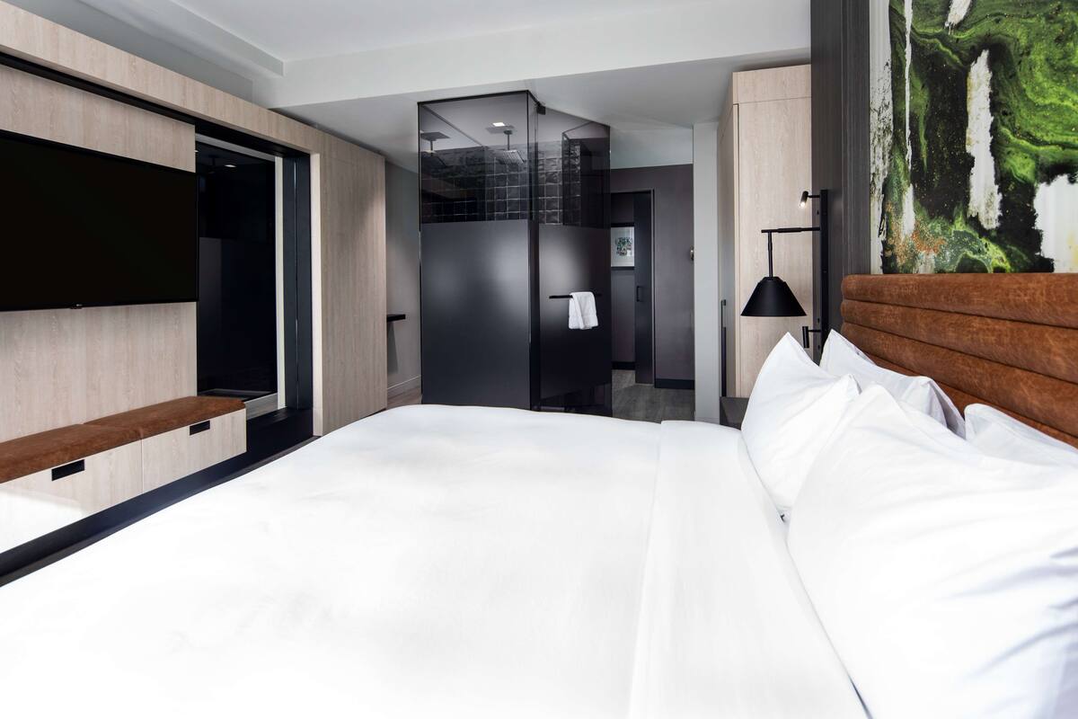 A modern hotel room with a large bed, a flat-screen TV, a wooden bench, and a painting on the wall.
