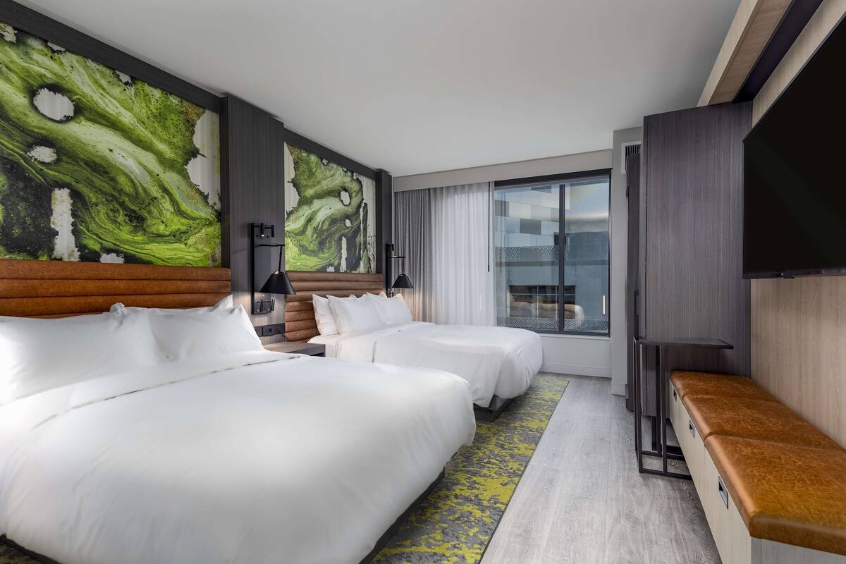 A hotel room with two beds, a large artwork on the wall, a flat-screen TV, and a large window with curtains.