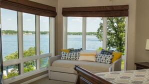 A bedroom with a large window overlooking a lake, a bed, and a sofa with patterned cushions.