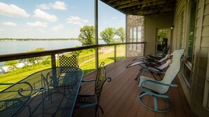 A covered deck with outdoor furniture, a view of a lake, and a stone building in the background.