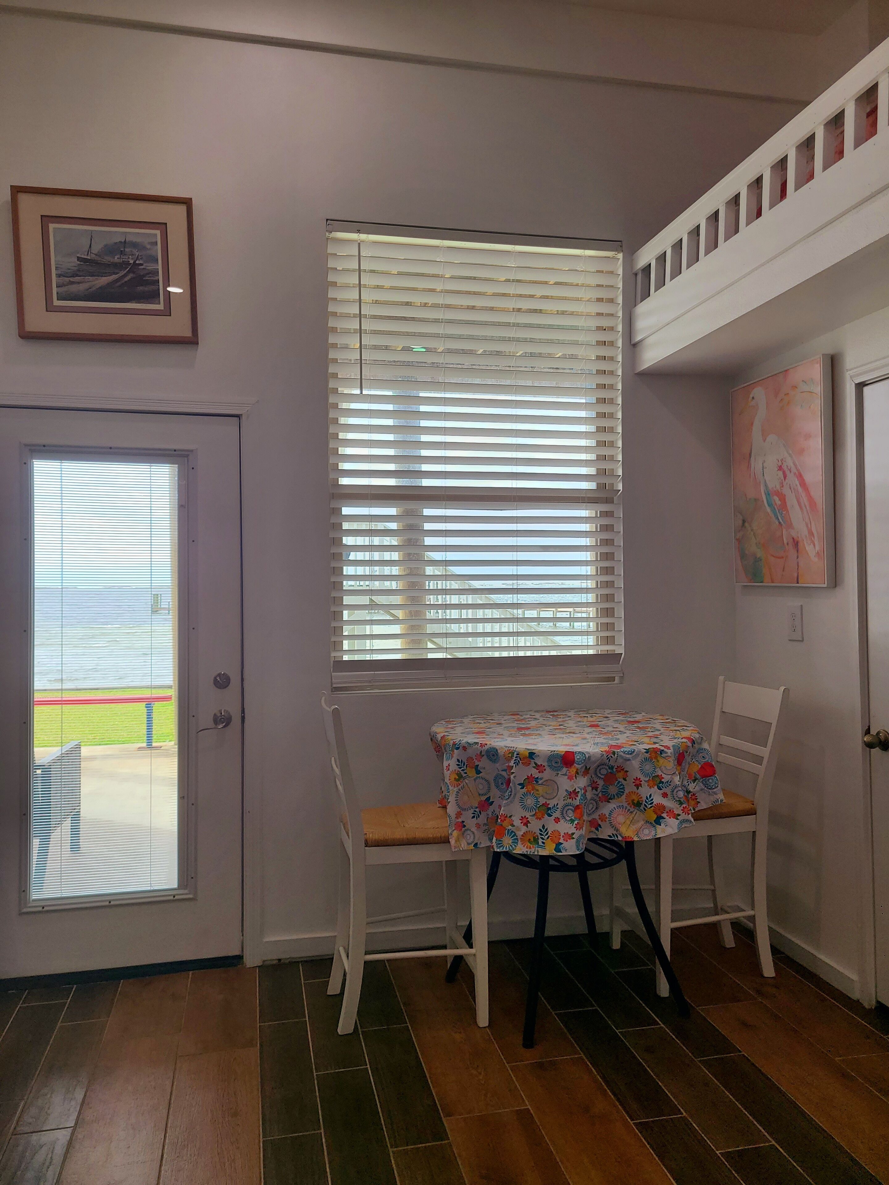 Une pièce comprenant une table à manger, des chaises, une fenêtre avec des stores, une porte et un tableau accroché au mur.