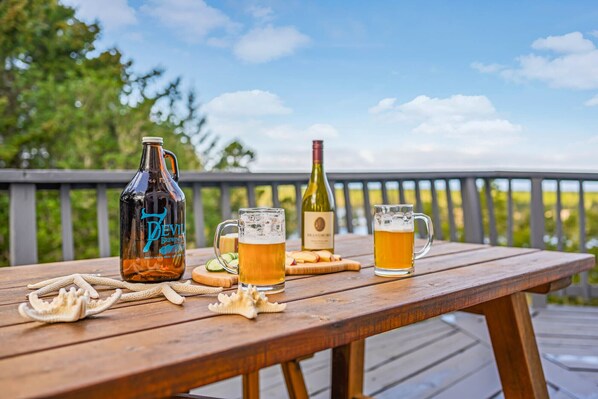 A wooden table with a beer bottle, two glasses of beer, a wine bottle, and some snacks on a deck.