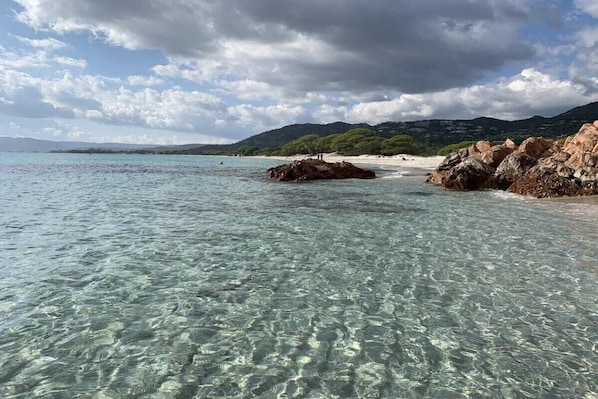 Uma praia com águas cristalinas, afloramentos rochosos e colinas ao longe.