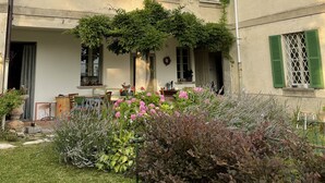 Ein mehrstöckiges Gebäude mit grünen Fensterläden, einer Veranda und einem Garten mit verschiedenen Pflanzen und Blumen.