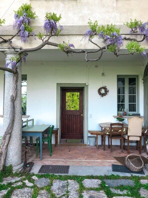 A covered patio with a wooden table and chairs, a door with a green mat, and purple wisteria hanging from the ceiling.