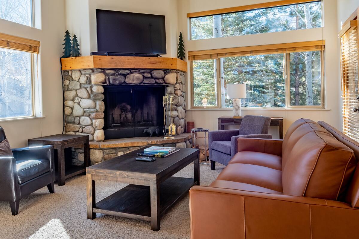 A cozy living room with a stone fireplace, leather furniture, and large windows offering a view of trees.