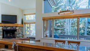 Une cuisine moderne dotée de plans de travail en granit, d’une cheminée en pierre, d’un téléviseur à écran plat et de grandes fenêtres offrant une vue sur les arbres.