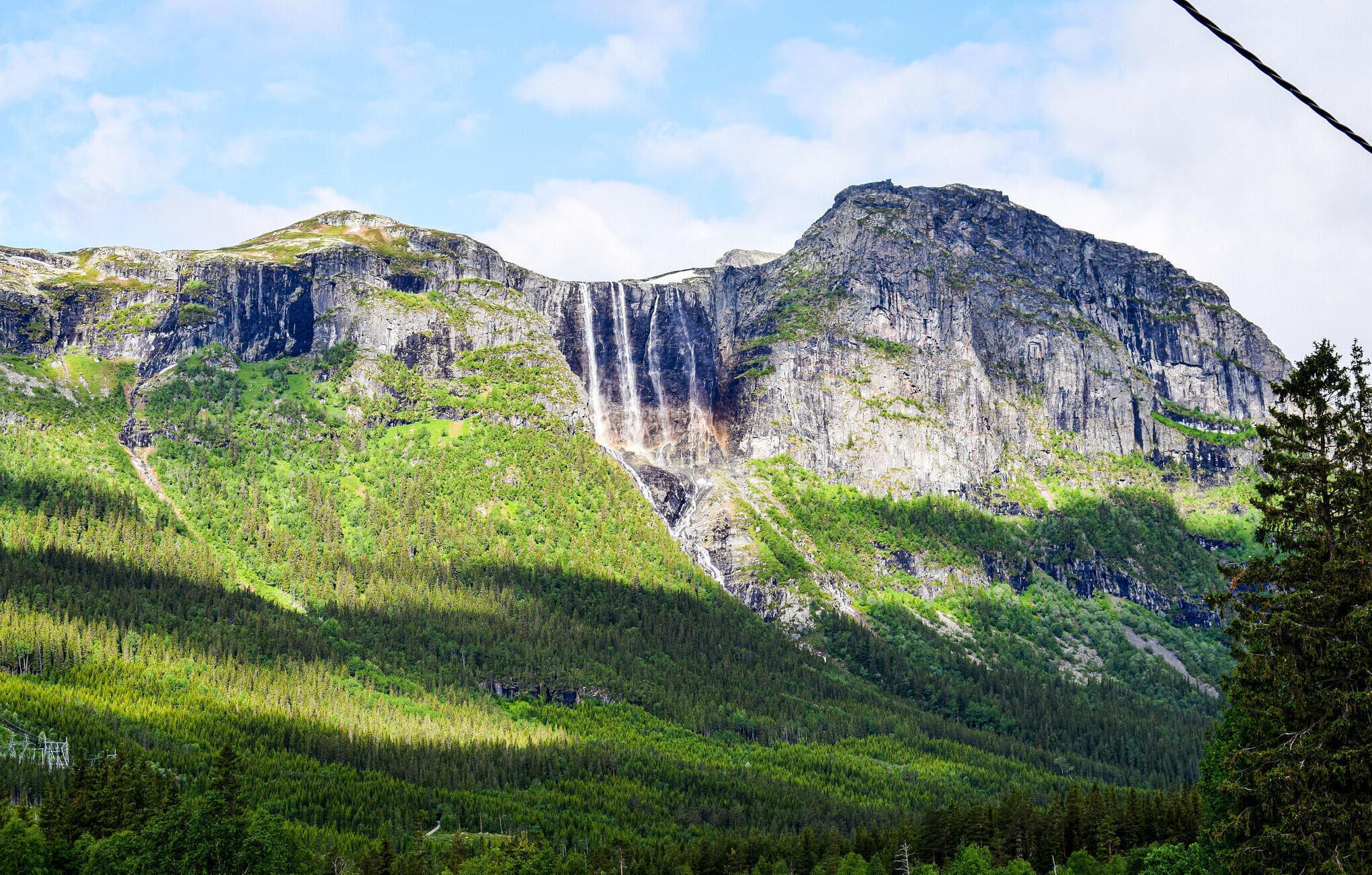 Een bergachtig landschap met een waterval, dicht bos en een heldere lucht.