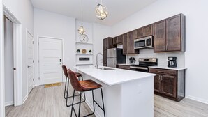 Une cuisine moderne avec un îlot central blanc, des meubles en bois foncé, des appareils électroménagers en acier inoxydable et des luminaires suspendus.