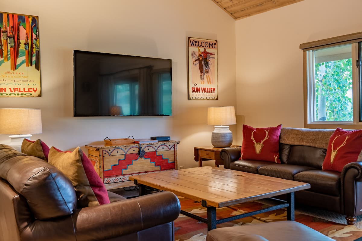 Una sala de estar con un sofá de cuero, una mesa de centro de madera y un televisor montado en la pared. Hay almohadones decorativos y un cartel en la pared.