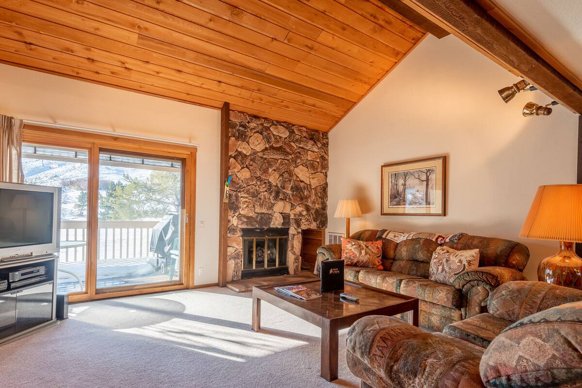 A cozy living room with a stone fireplace, a sofa, a television, and a large window overlooking a deck.