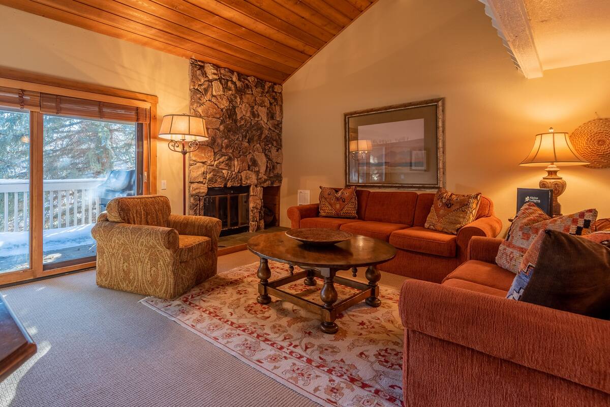 A cozy living room with a fireplace, a stone wall, and a large window overlooking a snowy deck.