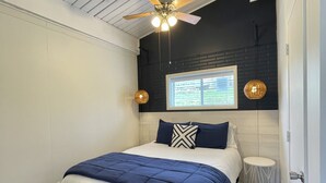 Un dormitorio con cama, ventilador de techo, ventana y una pequeña mesita de noche.