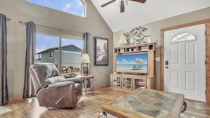 A spacious living room with a leather recliner, a television on a wooden stand, and a large window overlooking a house.