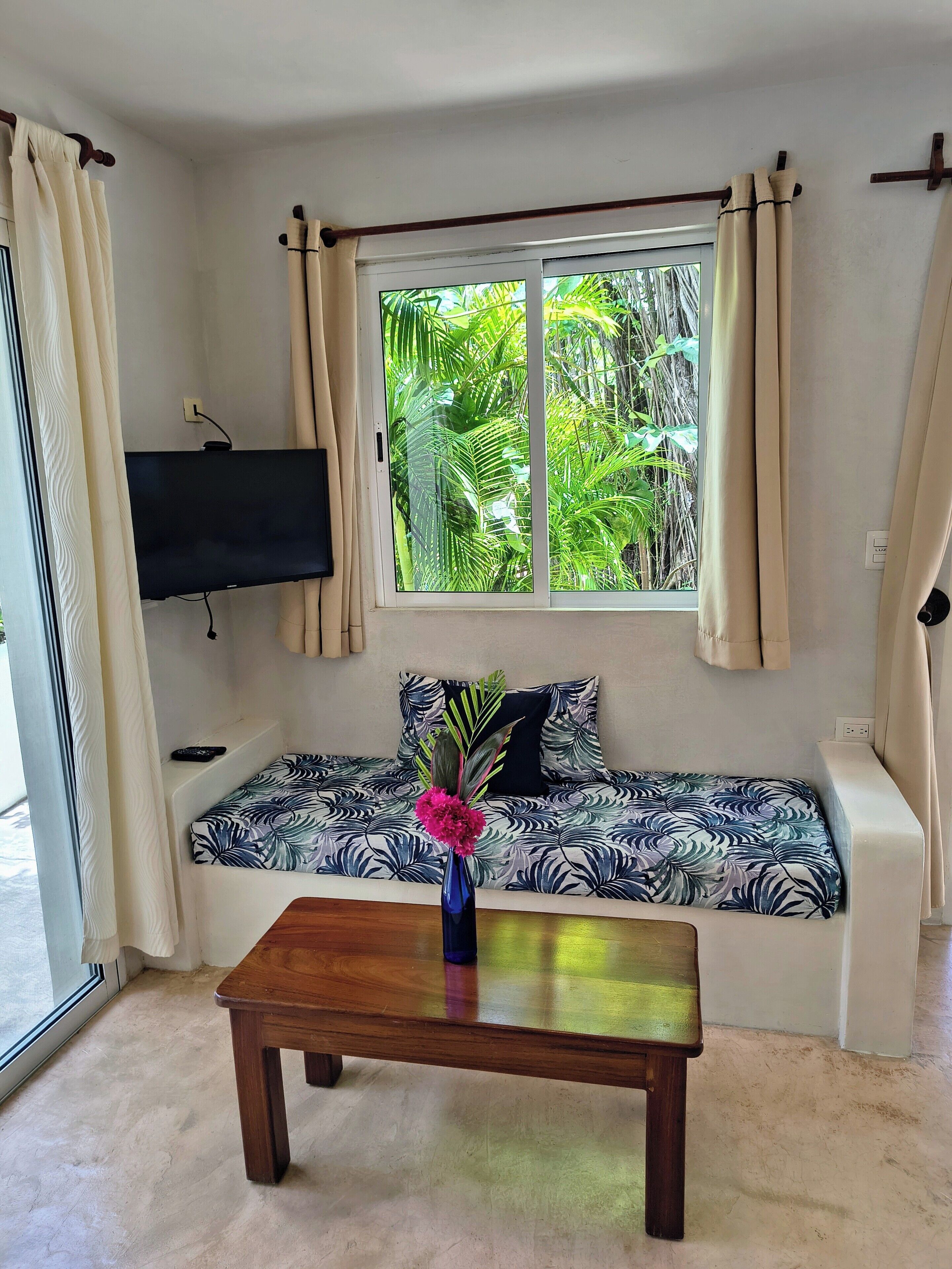 A living room with a sofa, a coffee table, a TV mounted on the wall, and a window with curtains.