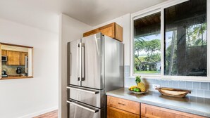 A kitchen with a stainless steel refrigerator, wooden cabinets, and a window with a view of greenery. - Keauhou Kona Surf & Racquet Club #1-103 (Kailua-Kona)