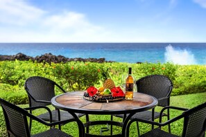 A round table with a woven placemat, two wicker chairs, a bottle, and a fruit platter with a pineapple, orange, and red flowers.