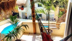 Une personne se détend dans un hamac sur un balcon donnant sur une piscine, agrémenté de plantes en pot, et offrant une vue sur un bâtiment doté d’un toit de chaume.