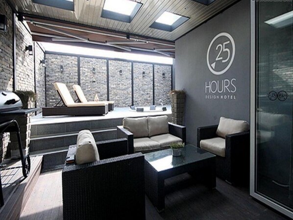 Um lobby de hotel moderno com uma parede de pedra, uma área de estar com sofás e uma mesa de centro, e uma porta de vidro com o número 25 e a inscrição "Hours Design Hotel".