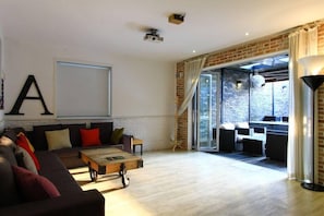 A spacious living area with a large sofa, a coffee table, and a fireplace. There is a large window with curtains, a brick wall, and a balcony with outdoor seating. - 25 Hours Hotel 2 (Busan)