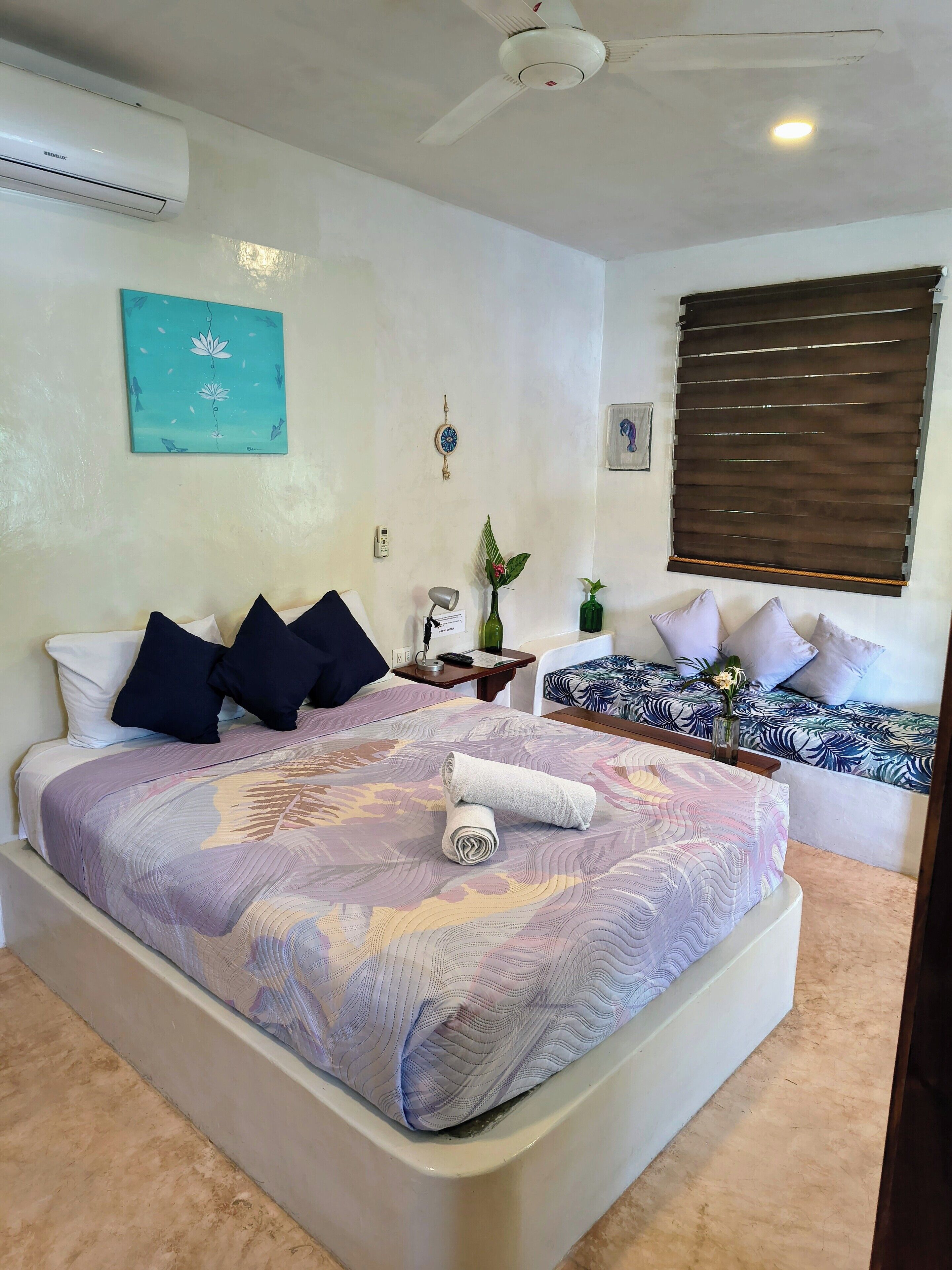 A hotel room with a large bed, a bedside table with a lamp, a small table with a vase, and a window with blinds.