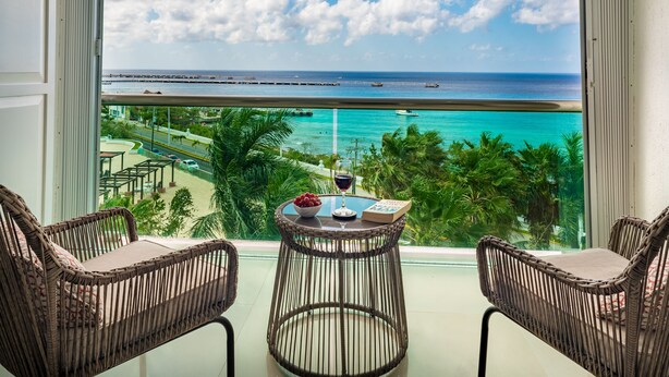 A balcony with wicker chairs, a small table with a glass of wine, and a view of the ocean and beach.