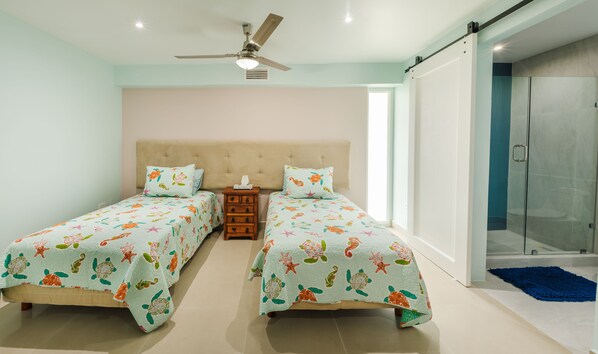 A bedroom with two beds, a ceiling fan, a nightstand, and a glass shower door.