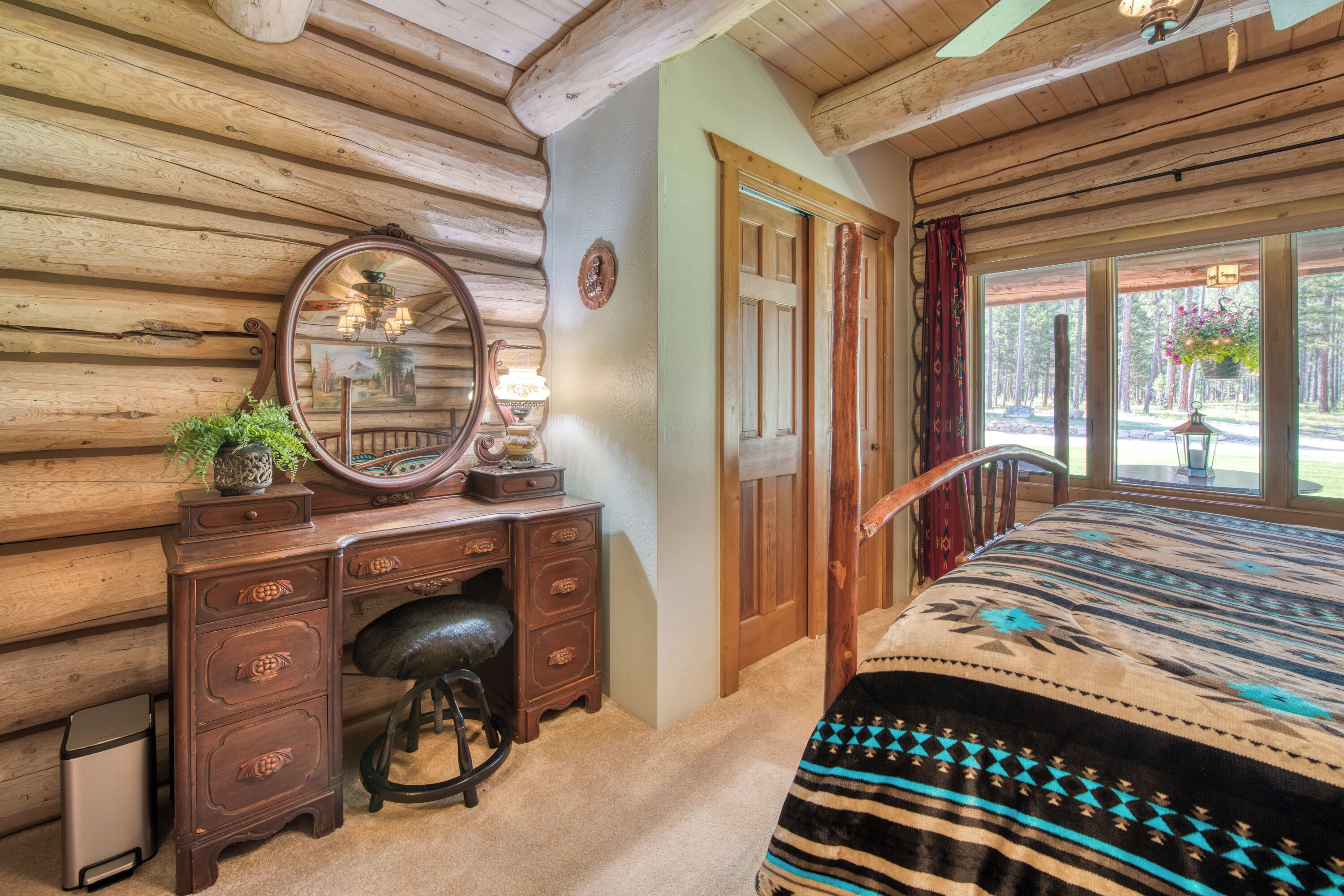 Quarto com teto de madeira, cama com manta estampada, cômoda de madeira com gavetas, espelho e janela com cortinas.
