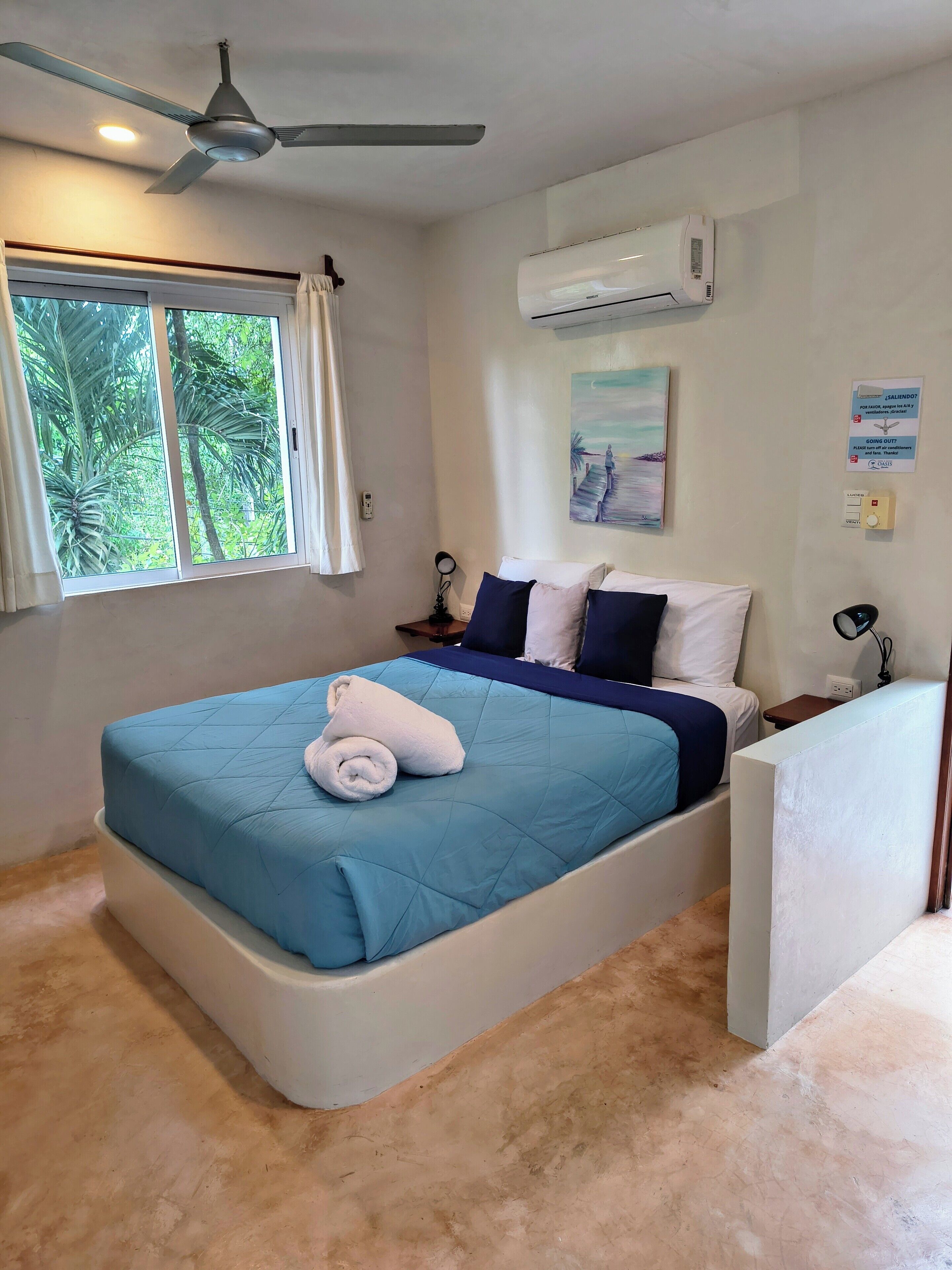 Un dormitorio con cama, ventilador de techo, aire acondicionado, ventana con cortinas y un cuadro en la pared.