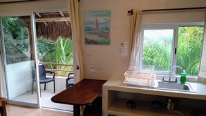 A room with a wooden table, a sink with a green soap dispenser, a trash bin, and a window with a view of greenery.