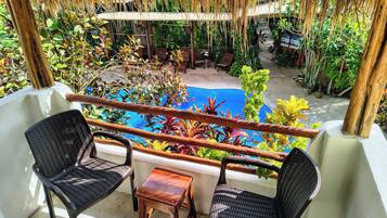 Un balcón con dos sillas de mimbre, una pequeña mesa de madera y una piscina rodeada de exuberante vegetación tropical.