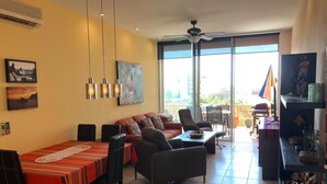 Uma sala de estar espaçosa com sofá, mesa de jantar e cadeiras. Há uma janela grande que oferece luz natural e vista para o exterior.