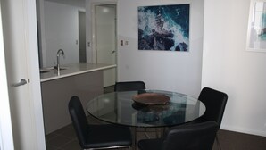 Uma área de refeições com uma mesa de vidro, quatro cadeiras, uma tigela de cobre, uma cozinha com uma pia e uma porta que dá para outro cômodo.