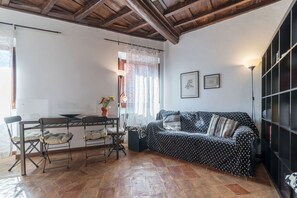 A cozy living room with a wooden ceiling, a black sofa with patterned cushions, a dining table with chairs, and a bookshelf.