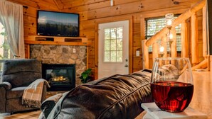 Ein gemütliches Cabin-Interieur mit Kamin aus Stein, einer Ledersofa und einem Fernseher, der über dem Kamin angebracht ist, sowie einem Glas Rotwein auf einem Tisch.