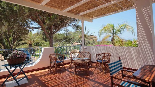 Patio esterno con mobili in vimini, un tavolo di legno e vista su alberi e un edificio sullo sfondo.