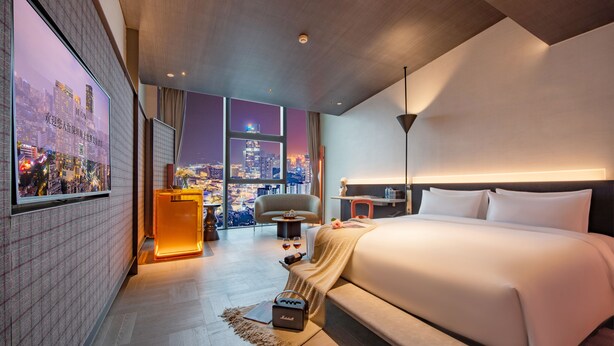 Een moderne hotelkamer met een groot bed, stadszicht, een bureau met stoel en een televisie aan de muur.