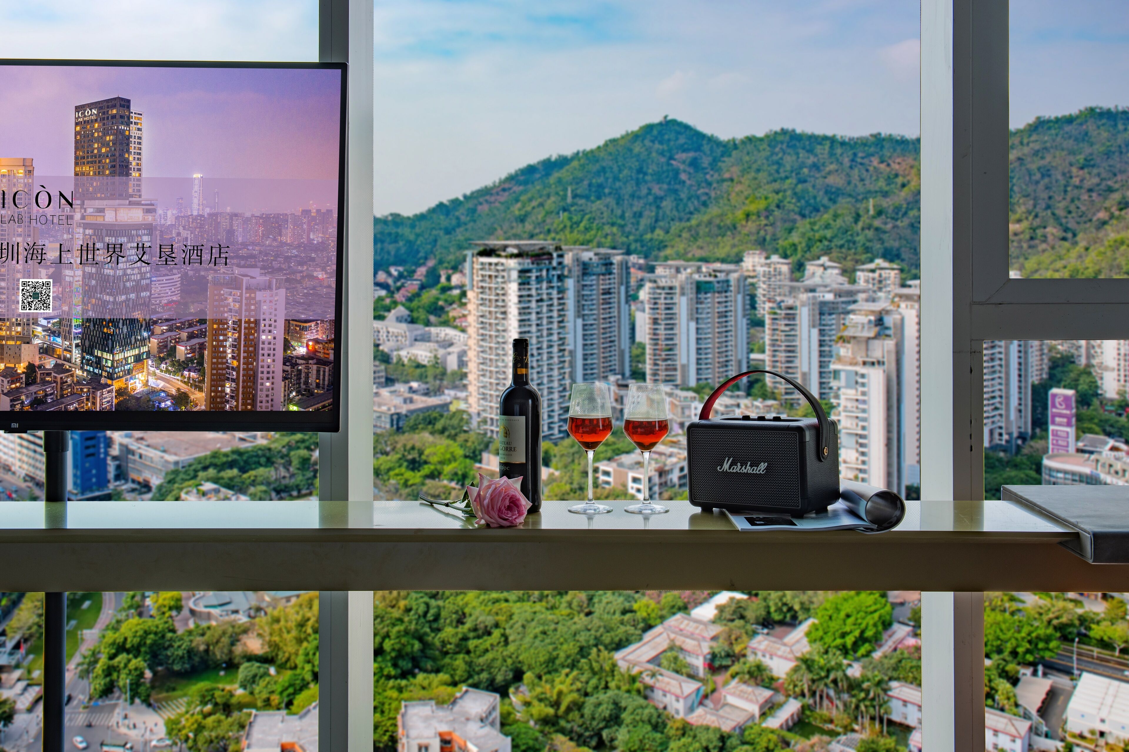 A cityscape view from a hotel room with a television displaying a cityscape, a bottle of wine, two wine glasses, and a portable speaker on a window sill.