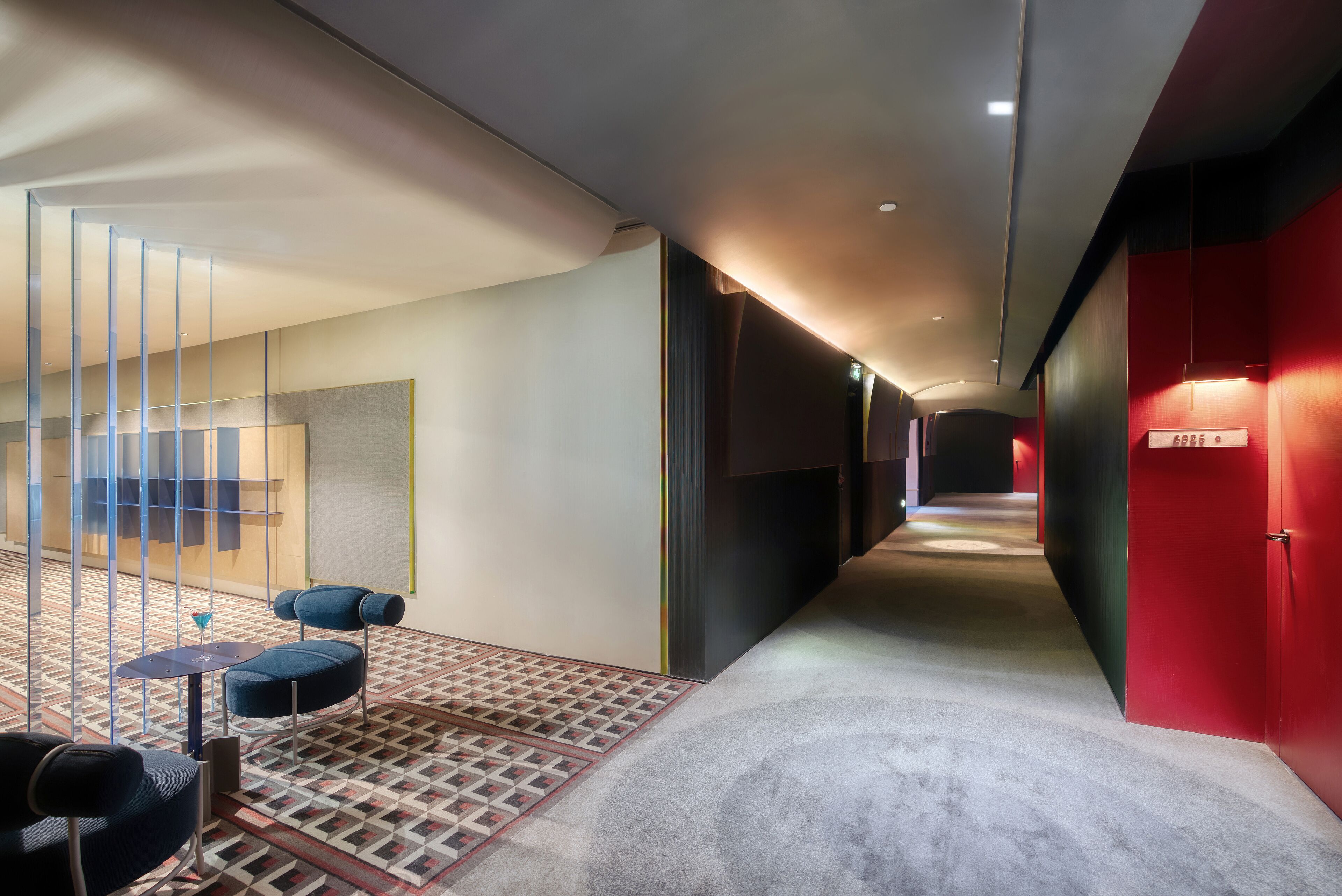 Un couloir d’hôtel moderne avec un tapis à motifs géométriques, un coin salon avec une petite table et des chaises, et un mur d’accent rouge.