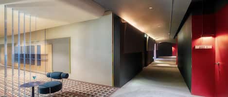 Un pasillo moderno de hotel con alfombra de diseño geométrico, un área de descanso con una pequeña mesa y sillas, y una pared de acento roja.
