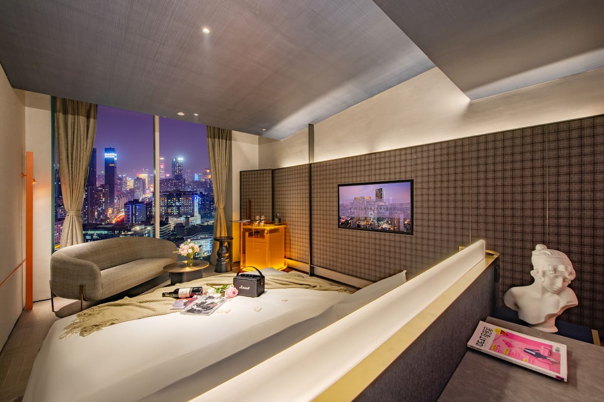 Habitación de hotel moderna con vista a la ciudad, un sofá, una mesita de noche con lámpara, un televisor y una escultura de busto.