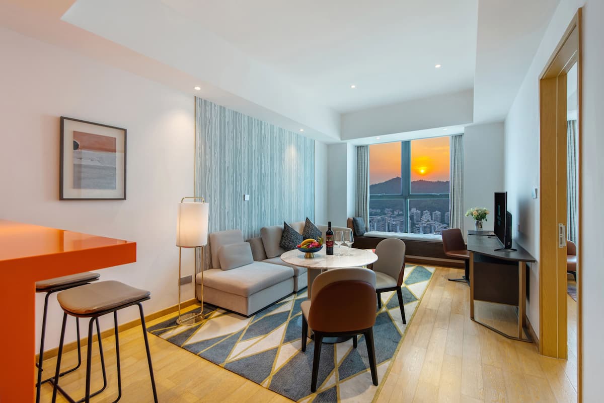 Habitación de hotel moderna con sofá, mesa de comedor y zona de bar. Cuenta con un ventanal grande con vistas a la ciudad y un atardecer.