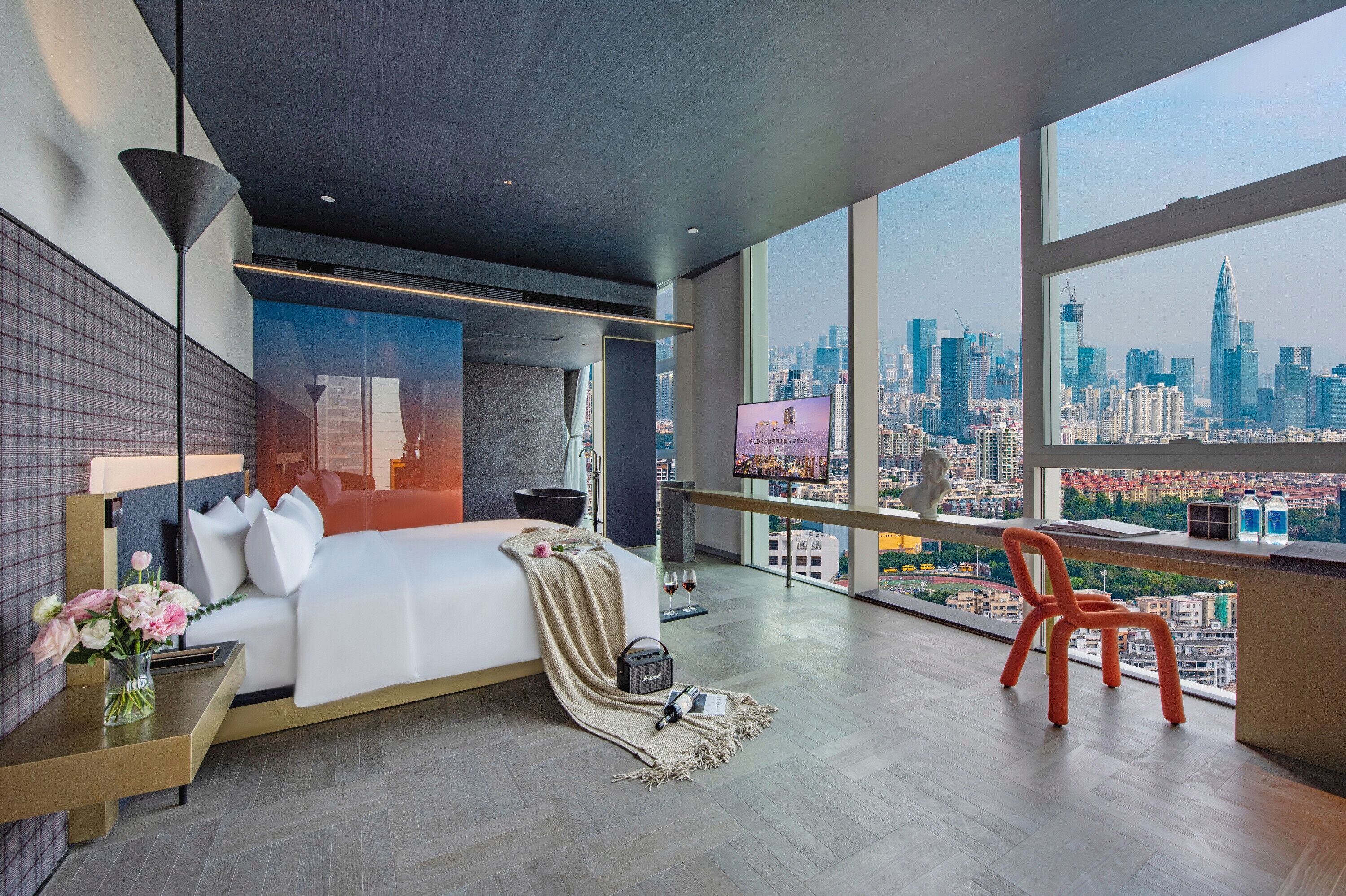 A modern hotel room with a large bed, a view of the city skyline, a balcony with a chair, and a flat-screen TV.