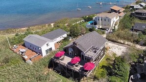 Een kustpand met een groot huis, een terras met parasols, een kleiner bijgebouw en uitzicht op zee met boten.