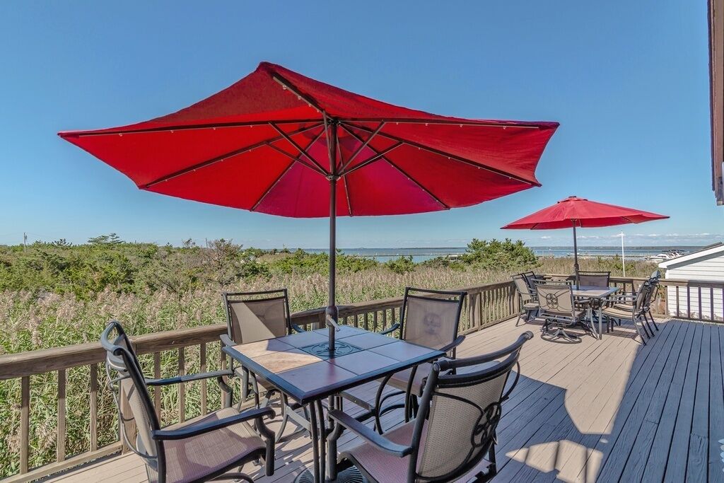 A deck with outdoor furniture and red umbrellas, overlooking a beach and ocean.