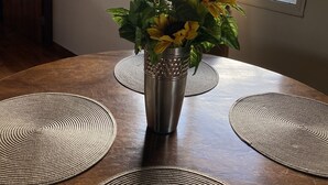 Una mesa de comedor con un jarrón metálico que contiene flores amarillas, rodeada de individuales tejidos, en una habitación con una ventana y un cuadro en la pared.
