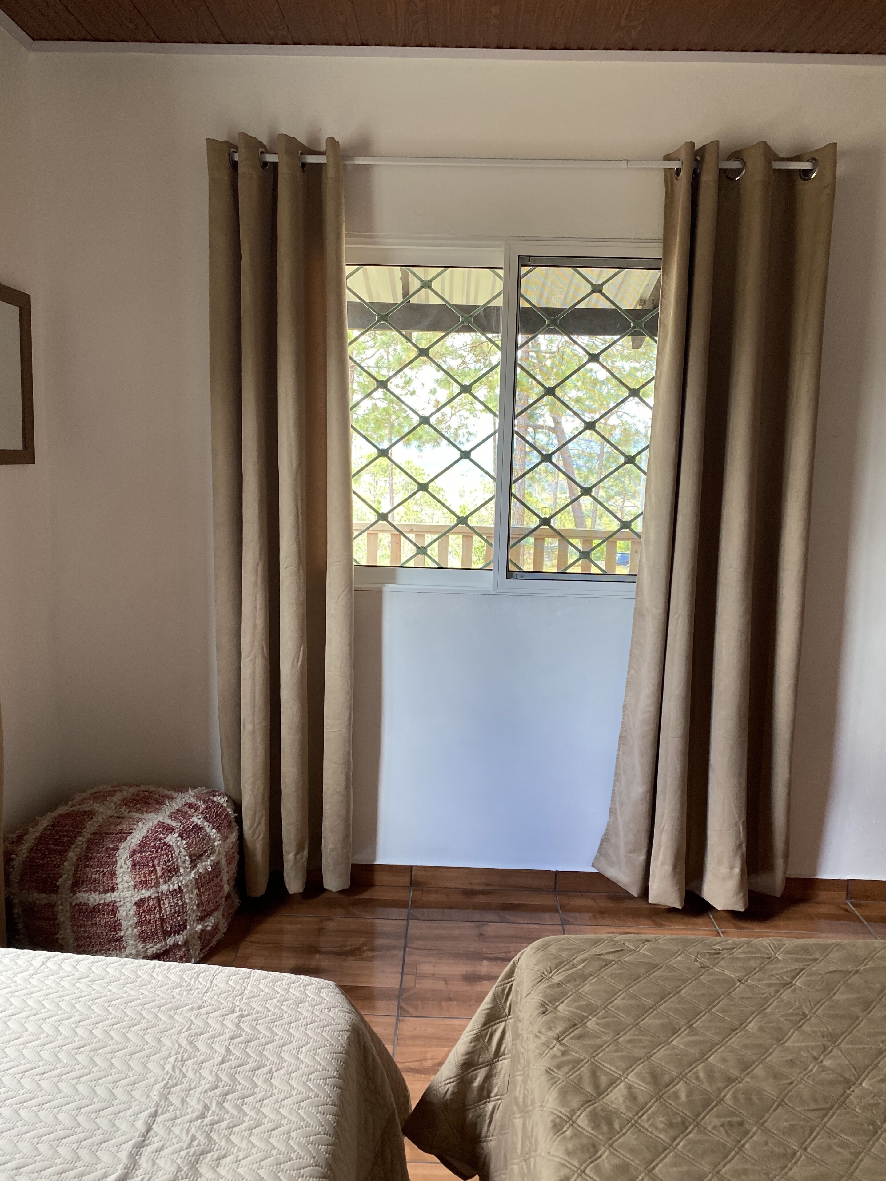 A room with two beds, beige curtains, a window with a view of trees, and a wooden ceiling.